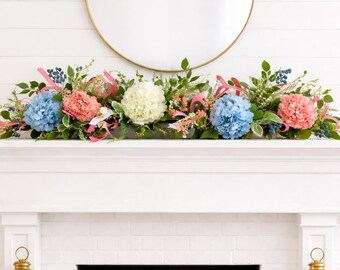 May include: A fireplace mantel decorated with a floral arrangement of blue, pink, and white hydrangeas, greenery, and pink ribbons. A round gold mirror hangs above the mantel. Gold lanterns flank the fireplace.