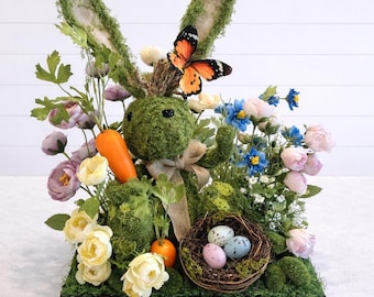 May include: An Easter decoration featuring a moss-covered bunny with large ears, surrounded by colourful flowers, a carrot, and a nest with three speckled eggs. A butterfly rests on the bunny's head. The arrangement is set on a wooden base.