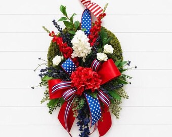 May include: A teardrop-shaped wreath featuring a green moss base adorned with red, white, and blue floral arrangements. The design includes red and white flowers, dark blue berries, and multiple ribbons with red, white, and blue star and stripe patterns.