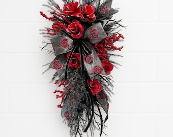 May include: A teardrop-shaped floral swag featuring deep red roses, clusters of small red berries, and dark black pine-like branches. The arrangement is accented with a large, textured grey ribbon and thin black velvet ribbons cascading downwards.