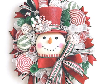 May include: A Christmas wreath featuring a snowman with a red top hat and scarf, surrounded by red and white striped sweets and green accents. The wreath has a large red bow and is ideal for festive home decor.