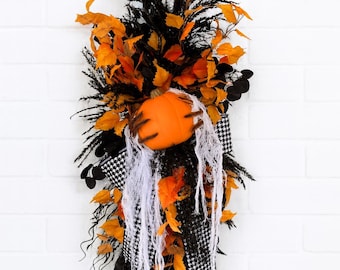 May include: A vertical Halloween door swag featuring a central orange pumpkin accented with dark brown skeletal hands. The arrangement includes black foliage, orange autumn leaves, white spider web material, and a black and white houndstooth ribbon.