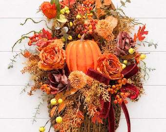 May include: A decorative wall hanging featuring a brown woven basket filled with an arrangement of orange pumpkins, artificial roses in shades of orange and deep red, dried foliage, yellow berries, and a dark red velvet ribbon with long tails.