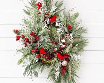 May include: A decorative wreath featuring green pine branches, frosted foliage, and brown pinecones. The arrangement is accented with clusters of bright red berries and three vibrant red cardinal bird figures nestled within the greenery.