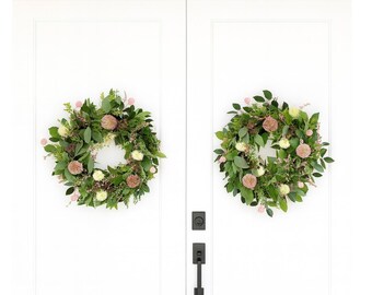 May include: Two circular wreaths made of green foliage, pink and cream flowers, adorn white doors. The wreaths have a natural, organic look. The doors feature black door handles and a lock.