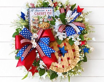 May include: A decorative wreath featuring a sign that reads All American CHERRY PIE, adorned with red, white, and blue ribbons, star-shaped accents, artificial white flowers, green foliage, and a fabric cherry pie cutout with red cherries.