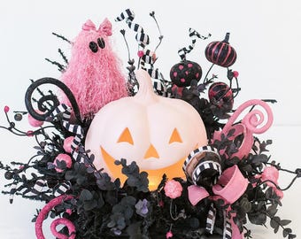 May include: A Halloween centrepiece featuring a light pink pumpkin with carved eyes and mouth, surrounded by black foliage, pink accents, and decorative elements. Includes a pink fuzzy monster and black and white striped ribbons.