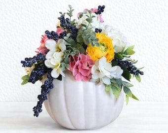 May include: A white, pumpkin-shaped ceramic vase filled with a colourful floral arrangement. The flowers include yellow, pink, white, and blue blooms, with green foliage. The vase has a ribbed design and sits on a white surface.