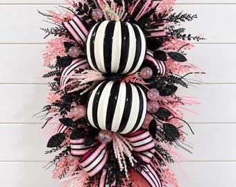 May include: A vertical wall hanging featuring two stacked black and white striped pumpkins centred on a pink wooden board. The arrangement is decorated with black glittery leaves, pink sprigs, iridescent spheres, and striped pink and black ribbons.