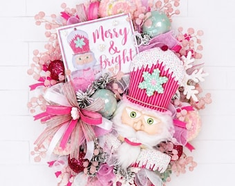 May include: A decorative wreath featuring a nutcracker figure with a pink hat, a sign reading Merry & Bright, pink berry sprays, white snowflakes, and assorted pink and mint green ornaments. The design incorporates layered ribbons and mesh.
