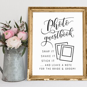 May include: A white sign with gold frame, with the text "Photo Guestbook" in black cursive font. The sign instructs guests to "Snap it, Shake it, Stick it... and leave a note for the bride & groom!"