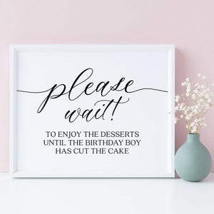 Puede incluir: Un letrero enmarcado en blanco con las palabras "please wait!" en una elegante escritura. Debajo, el texto dice "To enjoy the desserts until the birthday boy has cut the cake." Un pequeño jarrón azul claro contiene flores blancas.