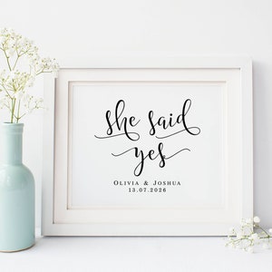 May include: White framed print with black calligraphy that reads "She said yes" with the names Olivia & Joshua and the date 13.07.2026 below. The print is sitting on a white surface with white flowers on either side.