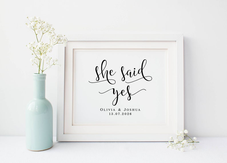 May include: White framed print with black calligraphy that reads "She said yes" with the names Olivia & Joshua and the date 13.07.2026 below. The print is sitting on a white surface with white flowers on either side.