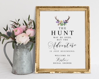 May include: A gold-framed sign with the text "The Hunt May Be Over, But The Adventure Is Just Beginning. Welcome To Karla's Bridal Shower."  The sign features a floral design with a deer silhouette.
