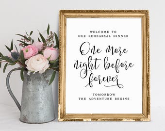 Pode incluir: Uma placa emoldurada com borda dourada exibe o texto "Bem-vindos ao nosso jantar de ensaio. Mais uma noite antes do para sempre. Amanhã a aventura começa." Um jarro de metal contém flores cor-de-rosa e brancas.