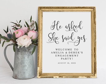 May include: A gold framed sign with the text "He asked She said yes" in black cursive font. Below the text is a welcome message for Amelia & Derek's engagement party on August 18, 2025.