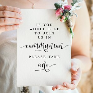 May include: A white card with black text that reads "If you would like to join us in communion please take one". The card is being held by a person wearing a white dress and a bouquet of pink flowers.