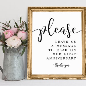 K&ouml;nnte beinhalten: Ein gerahmtes Schild mit goldfarbenem Rahmen und dem Wort "please" in eleganter schwarzer Schrift. Das Schild bittet die G&auml;ste, eine Nachricht f&uuml;r einen Jahrestag zu hinterlassen. Links steht ein Metallkrug mit rosa und wei&szlig;en Blumen.