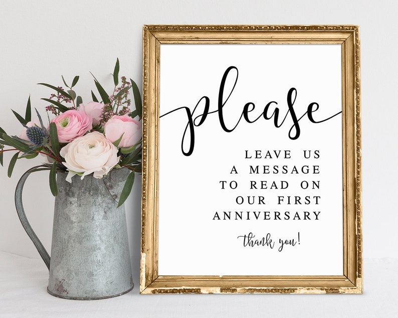 K&ouml;nnte beinhalten: Ein gerahmtes Schild mit goldfarbenem Rahmen und dem Wort "please" in eleganter schwarzer Schrift. Das Schild bittet die G&auml;ste, eine Nachricht f&uuml;r einen Jahrestag zu hinterlassen. Links steht ein Metallkrug mit rosa und wei&szlig;en Blumen.