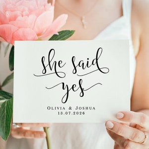 May include: A white card with black calligraphy that reads "She said yes" with the names Olivia & Joshua and the date 13.07.2026.