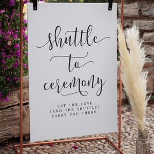 May include: A white sign with the words "Shuttle to Ceremony" in elegant script, held by a copper-colored metal frame. The sign also includes the text "Let the love (and the shuttle) carry you there." The sign is outdoors.