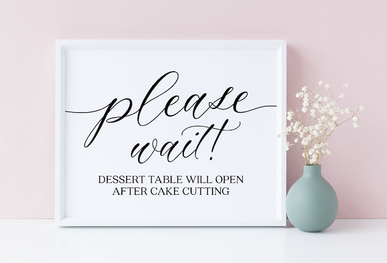 May include: A white framed sign with black script that reads "please wait!" and "DESSERT TABLE WILL OPEN AFTER CAKE CUTTING." A small vase with white flowers sits to the right of the sign. The background is a light pink.