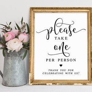 May include: A gold framed sign with white background and black text that reads "Please take one per person. Thank you for celebrating with us!" with a small black heart symbol.