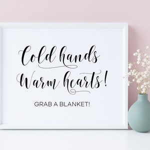 May include: White framed sign with black calligraphy text that reads "Cold hands warm hearts! Grab a blanket!" against a light pink background.