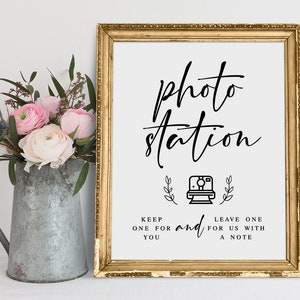 May include: A gold-framed sign with the text "photo station" in black script. The sign also includes a black and white illustration of an instant camera with leaves on either side. The text below the illustration reads "Keep one for you and leave one for us with a note."