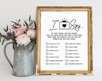 May include: A printable "I Spy" game card with a black and white camera icon and the text "I Spy" in a handwritten font. The card lists a variety of photo prompts for a wedding reception, such as "a photo of you", "a funny face", "a dance move", "a kiss", "a group photo", "a smiling bridesmaid", "a piece of cake", "a beautiful dress", "a new friend", "singing faces", "bride & groom", "making a speech", "a yummy treat", and "children having fun".