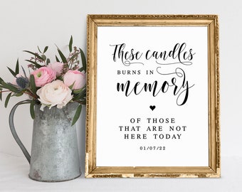 May include: A gold framed sign with the text "These candles burns in memory of those that are not here today 01/07/22". The sign is sitting on a white surface with a silver pitcher filled with pink flowers.