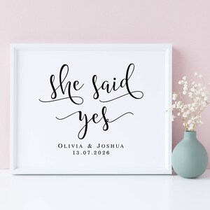 May include: White framed print with black calligraphy that reads "She said yes" with the names Olivia & Joshua and the date 13.07.2026 below. The print is on a white surface with a vase of white flowers in the background.