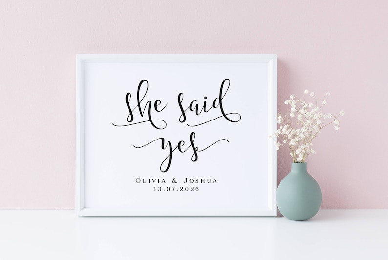 May include: White framed print with black calligraphy that reads "She said yes" with the names Olivia & Joshua and the date 13.07.2026 below. The print is on a white surface with a vase of white flowers in the background.