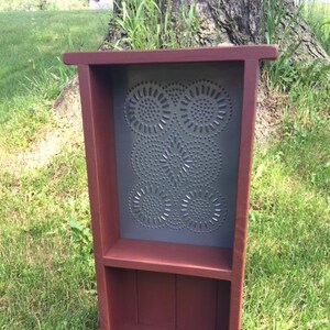 May include: A wooden shelf with two shelves and a tin panel with a decorative pattern. The shelf is painted a deep red color and the tin panel is a dark gray color.