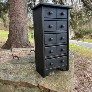 May include: A small, black wooden cabinet with six drawers, each with a round knob. The cabinet is painted black and sits on a stone surface. The cabinet has a simple, traditional design, suitable for storage.