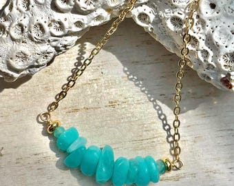 May include: A gold-colored chain necklace featuring a row of turquoise-colored, irregular-shaped stones. The stones are strung together, creating a curved design. The necklace is displayed on a light-colored, textured surface.