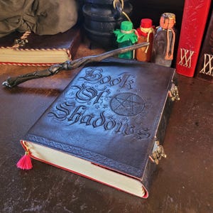 May include: A dark brown leather-bound book titled Book of Shadows with an embossed pentagram on the cover, featuring metal latches, a pink tassel bookmark, and a dark wooden wand resting on top, accompanied by small glass potion bottles.