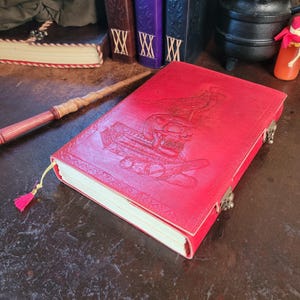 May include: A bright red leather-bound journal featuring an embossed design of a skull, a bird, and a scroll on the front cover. The book includes a metal latch closure, a yellow ribbon bookmark with a pink tassel, and off-white pages.
