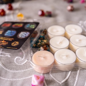 May include: A clear plastic container holding eight white tea light candles and one peach-colored candle. The container also holds a set of dice and a black label with colorful text. Other dice are scattered around.