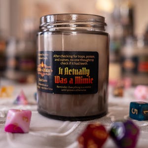 May include: A brown candle in a glass jar with a silver lid. The label reads "It Actually Was a Mimic." Colorful dice surround the candle. The candle is a dual scent with a burn time of 40-50 hours.