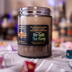 May include: A brown candle in a glass jar with a black label that reads "We Split The Party." The candle is surrounded by colourful dice, suggesting a tabletop gaming theme. The text on the label includes a warning about potential death scenes.