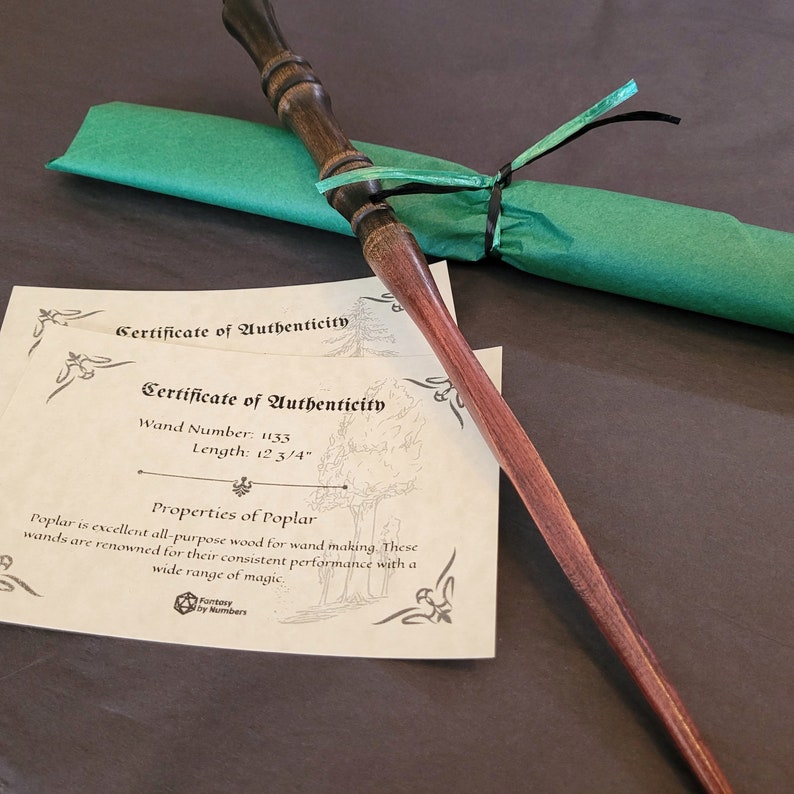 May include: A handcrafted wooden wand with a dark brown handle and a reddish-brown shaft. The wand is 12 3/4 inches long and is accompanied by a green paper sleeve tied with black and green ribbons. Two certificates of authenticity are also present.