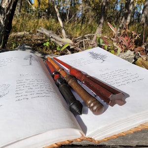May include: Four handcrafted wooden wands in varying shades of brown and red rest on an open book with handwritten text and tree illustrations. The wands have unique shapes and finishes, suggesting a magical theme.