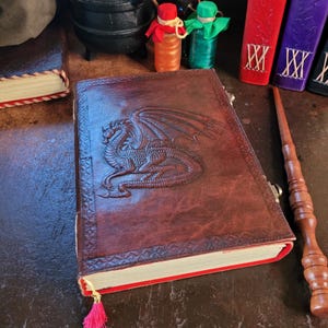 May include: A dark brown leather-bound journal featuring an embossed dragon design on the front cover, a red fabric bookmark with a tassel, and a matching wooden wand placed alongside it. The journal has a textured border and cream-coloured pages.