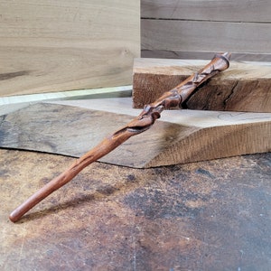 Puede incluir: Una varita de madera con un diseño único e intrincado. La varita está tallada con patrones en espiral y tiene un color marrón natural.