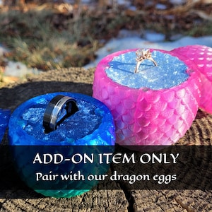 May include: Two dragon egg-shaped ring boxes, one blue and one pink, with a ring inside each. The text "ADD-ON ITEM ONLY Pair with our dragon eggs" is at the bottom of the image.