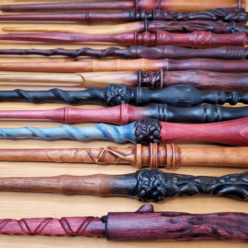May include: A collection of handcrafted wooden wands in various colors and designs. The wands feature intricate carvings, twisted handles, and a range of hues from dark brown and black to red and blue. Each wand has a unique shape and texture.