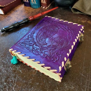 May include: A purple leather journal featuring an embossed raven design and Celtic knotwork. The journal has a stitched binding, brass clasps, and a teal tassel. A wooden wand is nearby.