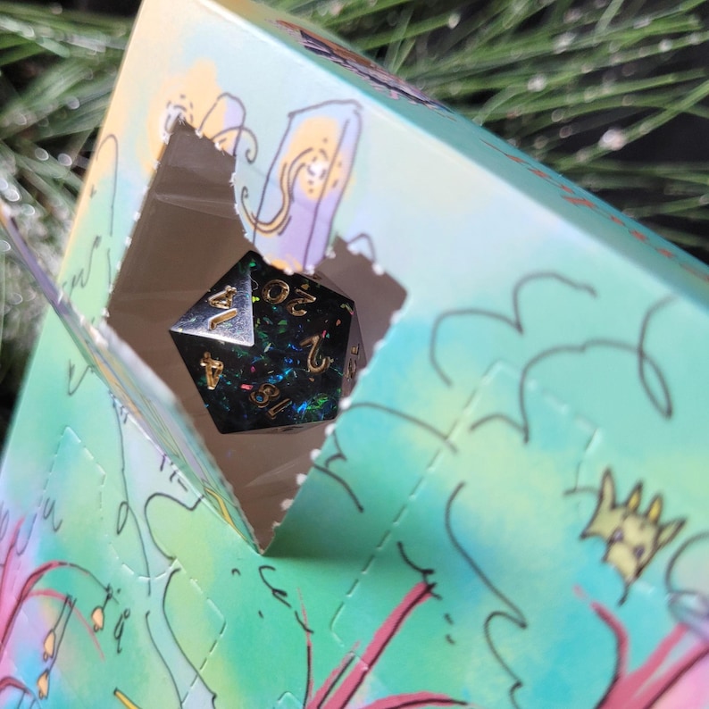 May include: A twenty-sided die with a dark, glittery finish and gold-colored numbers is visible through a square cutout in a light green and blue cardboard box featuring whimsical line-drawn illustrations of plants and creatures.